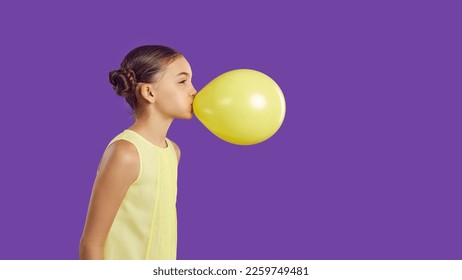 Cheerful funny little girl inflates yellow balloon isolated on vivid purple background. Profile portrait of cute preteen girl who inflates balloon near copy space. Children's holidays concept. Banner.