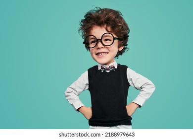 Cheerful funny little kid with dark wavy hair in classy clothes and round glasses, smiling brightly and looking away while standing against turquoise background with hands on waist - Shutterstock ID 2188979643