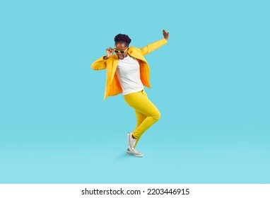 Cheerful funny woman in good mood dancing cheerfully isolated on light blue background. Young stylish African American woman in yellow casual suit, t-shirt, sneakers and sunglasses. Full length. - Powered by Shutterstock