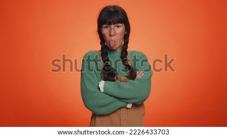 Cheerful funny woman in green sweater showing tongue making faces at camera, fooling around, joking, aping with silly face teasing. Young nerd adult girl isolated alone on orange studio background
