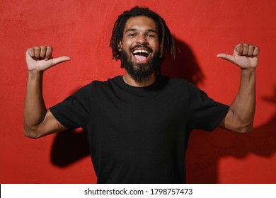 Cheerful funny young african american man with dreadlocks 20s wearing black casual t-shirt posing pointing thumbs on himself looking camera isolated on bright red color background studio portrait