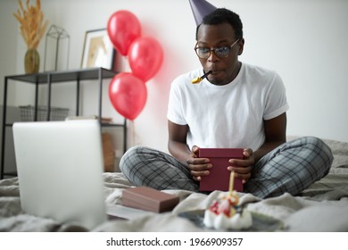 Cheerful Funny Young Afro American Male Celebrating His Birthday Online, Sitting On Bed With Cake And Presents, Blowing Party Horn, Having Video Conference Party With Friends And Family Using Laptop