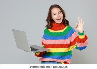 Cheerful funny young brunette woman in casual colorful knitted sweater standing showing OK gesture working on laptop pc computer looking camera isolated on grey colour background, studio portrait