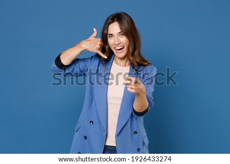 Cheerful funny young brunette woman 20s wearing basic jacket doing phone gesture like says call me back pointing index finger on camera isolated on bright blue colour background, studio portrait