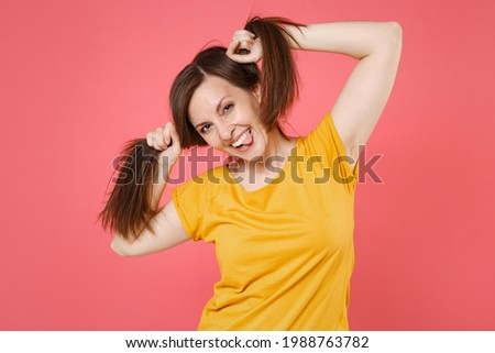 Cheerful funny young brunette woman 20s in yellow casual t-shirt posing hold hair like ponytails showing making stick tongue out sign, fooling around isolated on pink color background studio portrait