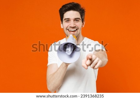 Cheerful funny young man 20s wearing basic casual empty blank white t-shirt standing screaming in megaphone pointing index finger on camera isolated on bright orange colour background studio portrait