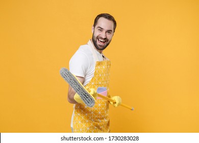 Cheerful funny young man househusband in apron rubber gloves hold in hands broom while doing housework isolated on yellow wall background studio portrait. Housekeeping concept. Looking camera