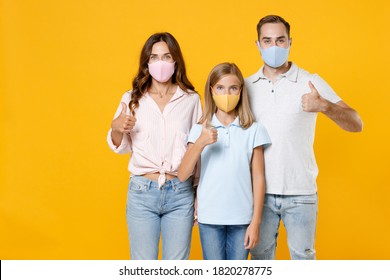 Cheerful Funny Young Parents Mom Dad With Child Kid Daughter Teen Girl In T-shirts Face Mask To Safe From Coronavirus Virus Covid-19 Showing Thumb Up Isolated On Yellow Background. Family Day Concept