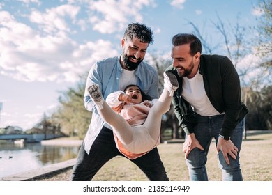 Cheerful Gay Couple Playing With Their Daughter In The Park. Father Swings His Little Girl In The Air, Who Laughs Out Loud.