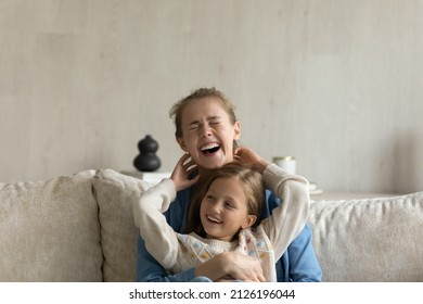Cheerful giggling kid and young mom hugging, laughing, having fun. Mother holding in arms, cuddling, tickling cute daughter child on couch at home. Childcare, family, motherhood concept