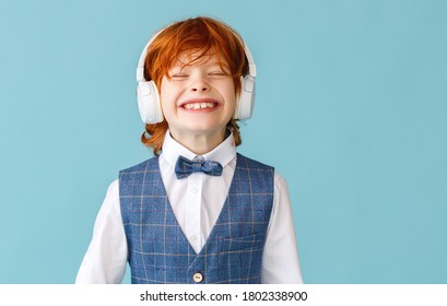 Cheerful Ginger Boy In School Uniform  With Closed Eyes Smiling While Listening To Music In Headphones Against Blue Background
