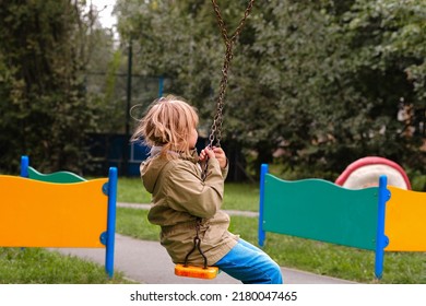 A Cheerful Girl Of 10 Years Old Is Swinging On A Swing In The Playground. A Child Plays Outside In Early Spring In The Yard.