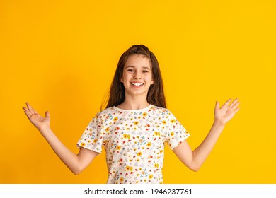 Cheerful Girl About 10 Years Old In A Colorful T-shirt Spreads Her Arms To The Sides And A Happy Smile On A Yellow Background. A Happy Child Enjoys Success And Victory. Discounts And Sales