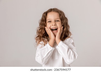 Cheerful girl about 5-6 years old in white shirt holding her hands near her face and happy smile on white background. happy child enjoys success and victory. Discounts and sales. Advertising concept