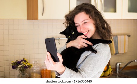 Cheerful Girl With Black Cat Using Mobile Phone For Video Call, Gesturing Hi To Friends, Relatives Or Parents. Smiling Young Woman Making Selfie, Video At Kitchen Home. Social Distancing, Isolation