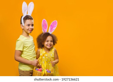 A Cheerful Girl And Boy With Rabbit Ears On Her Head With A Basket Of Colored Eggs In Her Hands On A Yellow Background. They Looks Away. Funny Happy Baby. Covid Easter To The Child.