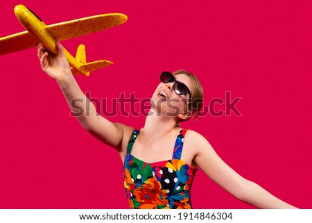 A cheerful girl in bright clothes and in sunny hands holds a yellow plane on a red background and dreams of a trip. Travel and tour with advertising space. High quality photo