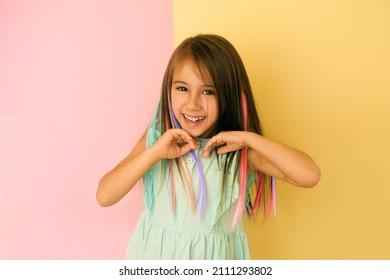 Cheerful girl with bright multicolored strands of hair laughs merrily looking at the cameras and straightening her hair with her hands. Child on a yellow-pink studio background. Stylish hair coloring