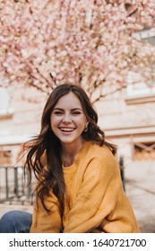 Cheerful Girl In Cashmere Sweater Laughs Against Backdrop Of Blossoming Sakura. Portrait Of Woman In Yellow Hoodie In City In Spring