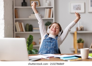 Cheerful girl in casual clothes and headphones raising clenched fists and smiling with closed eyes while celebrating success at end of online lesson at home