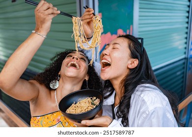 Cheerful girl friends- of Afro-Latin, and  Asian ethnicity, enjoying eating noodles outdoors together. They are smiling brightly while capturing a fun and joyful moment.