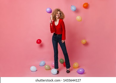 Cheerful girl with glass of champagne posing on pink background among balloons. Snapshot of curly woman in red top and denim pants