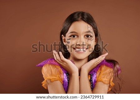 cheerful girl in Halloween costume and spiderweb makeup gesturing on brown backdrop, trick or treat
