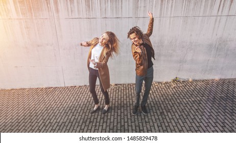 Cheerful Girl and Happy Young Man with Long Hair are Actively Dancing on a Street next to an Urban Concrete Wall. They Wear Brown Leather Jacket and Coat. Shot Took from Above on a Sunny Day.
