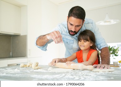 Cheerful Girl And Her Dad Kneading And Rolling Dough On Kitchen Table With Flour Messy. Father Teaching Daughter To Bake. Family Cooking Concept