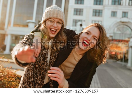 Cheerful girl in knitted hat walking with friend in city. Pleased ladies having fun in autumn.