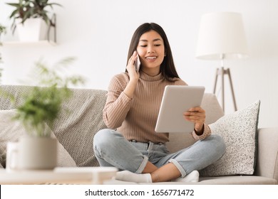 Cheerful girl making order by phone using digital tablet, sitting on sofa at home