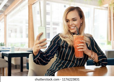 Cheerful girl making a selfie while sitting in a modern cafe and drinking a colorful drink with windows in the background. Perfect smile. - Shutterstock ID 1176223831