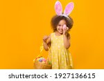 A cheerful girl with rabbit ears on her head with a basket of colored eggs in her hands on a yelow background. Funny crazy happy baby. Easter to the child.