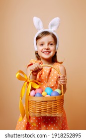 A Cheerful Girl With Rabbit Ears On Her Head And A Protective Mask With A Basket Of Colored Eggs In Her Hands On A Beige Background. Funny Crazy Happy Baby. Covid Easter To The Child.