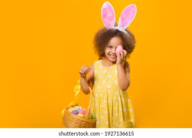 A cheerful girl with rabbit ears on her head with a basket of colored eggs in her hands on a yelow background. Funny crazy happy baby. Easter to the child. - Shutterstock ID 1927136615