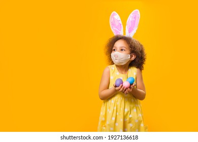 A Cheerful Girl With Rabbit Ears On Her Head And A Protective Mask With A Basket Of Colored Eggs In Her Hands On A Yellow Background. She Looks Away. Funny Happy Baby. Covid Easter To The Child.