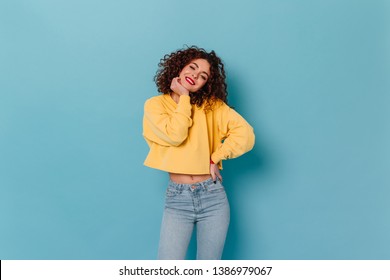 Cheerful girl with red lips and snow-white smile looks into camera on blue background. Portrait of cute curly lady in yellow top with long sleeves and jeans