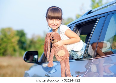 Cheerful girl is standing in car and smiling. She is pushing her body through window outside. The girl is holding toy and looking forward playfully