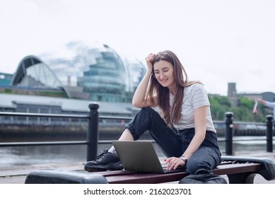A cheerful girl works with a laptop on the embankment sitting on a bench. The concept of an open-air office. Freelancer at work. Newcastle, The Sage Gateshead, North East England.