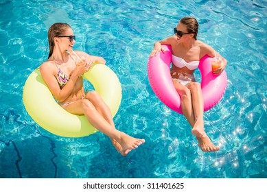 Cheerful girlfriends are having fun in swimming pool and drinking cocktail.