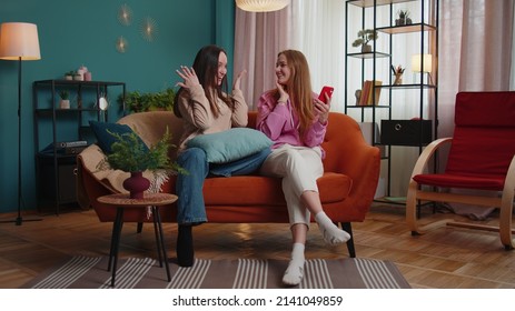 Cheerful Girls Friends Siblings Celebrate Success Win Scream Rejoices While Doing Online Shopping On Smartphone. Happy Two Female Women Couple Family On Sofa At Home Spend Leisure Time On Mobile Phone