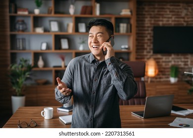 Cheerful Glad Successful Handsome Mature Chinese Male Calling By Phone, Talking To Client In Home Office Interior. Businessman Have Business Conversation Remote And Great News During Covid-19 Pandemic