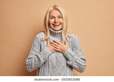 Cheerful good looking middle aged woman keeps hands pressed on chest smiles broadly and expresses positive emotions dressed in winter jumper happy to hear compliment isolated over beige background - Powered by Shutterstock