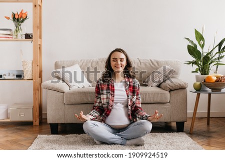 Cheerful good-humored pregnant woman meditating on beige soft carpet. Pretty brunette mother smiles in living room.