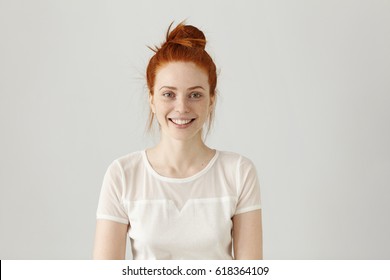 Cheerful gorgeous young woman wearing her ginger hair in knot smiling happily while receiving some positive news. Pretty girl dressed in white blouse looking at camera with excited joyful smile