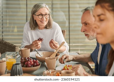 Cheerful grandmother in eyeglasses holding grape and looking at her family