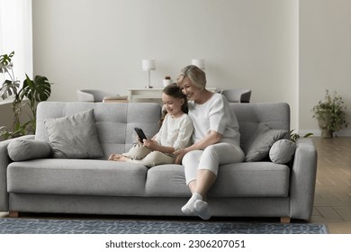 Cheerful grandmother and granddaughter sit on couch, use cellphone together, play on-line games, communicate with relatives through video call. Multi-generational family spend pastime with modern tech - Powered by Shutterstock