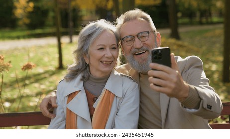 Cheerful gray-haired wife husband using smartphone online communication on bench in autumn park nature old grandparents senior couple mature family happy woman man talk with mobile phone outdoors city - Powered by Shutterstock