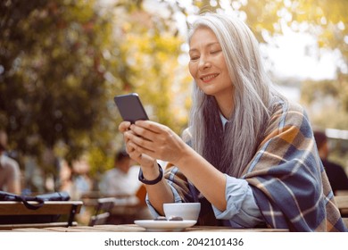 Cheerful grey haired senior Asian woman writes sms on smartphone sitting at table on outdoors cafe terrace on autumn day