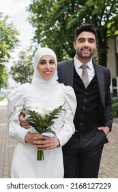 cheerful groom posing with hand in pocket near muslim bride in hijab with wedding bouquet